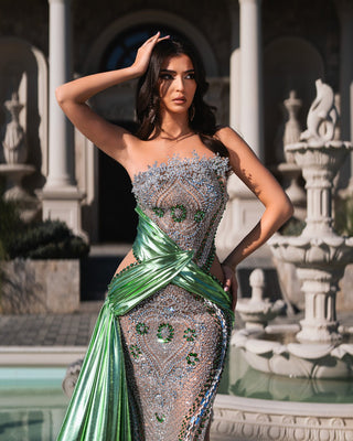 Elegant woman in a green crystal-embellished gown posing by a fountain.