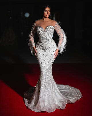 A glamorous bridal gown with a mermaid silhouette, crystal embellishments, sheer sleeves adorned with feathers, and a dramatic train.