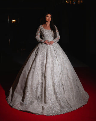 A luxurious ball gown with a fitted bodice, long sheer sleeves adorned with lace and beads, and a voluminous skirt embellished with shimmering sequins and metallic threads in soft metallic silver.