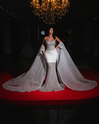 A sparkling strapless mermaid-style bridal gown with a crystal-embellished bodice and a flowing sheer cape-like train adorned with delicate glittering details.